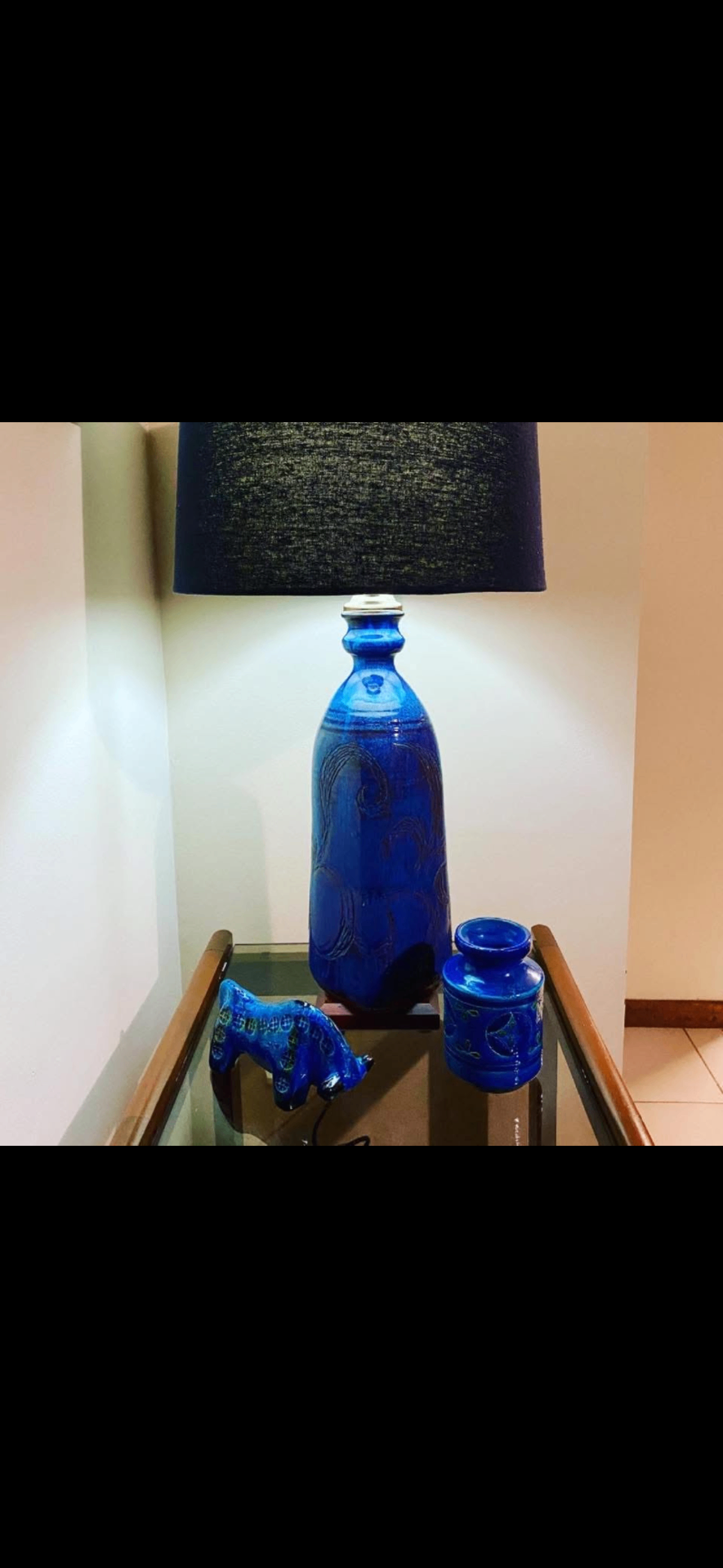 Tall cobalt Bitossi-style bottle lamp with a dark linen shade on a glass-top side table — a quiet corner detail.