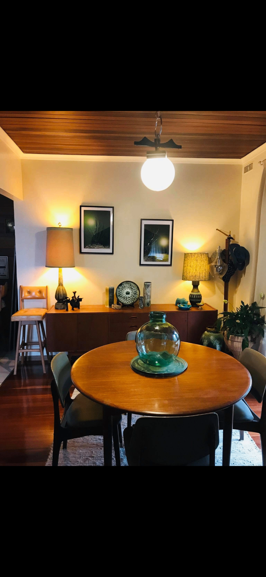 Mid-century dining room at dusk — two ceramic table lamps glowing on a teak sideboard beneath a timber-lined ceiling and a milk-glass pendant globe.